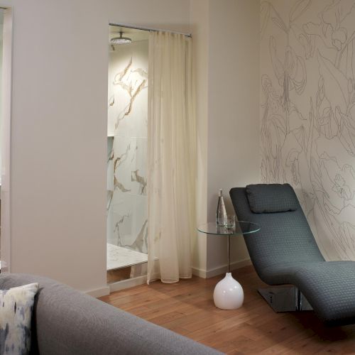 A modern, stylish living room with a black chaise lounge, a tall floor lamp, a patterned wall, and a doorway to a glass-walled shower area; stylish and serene.