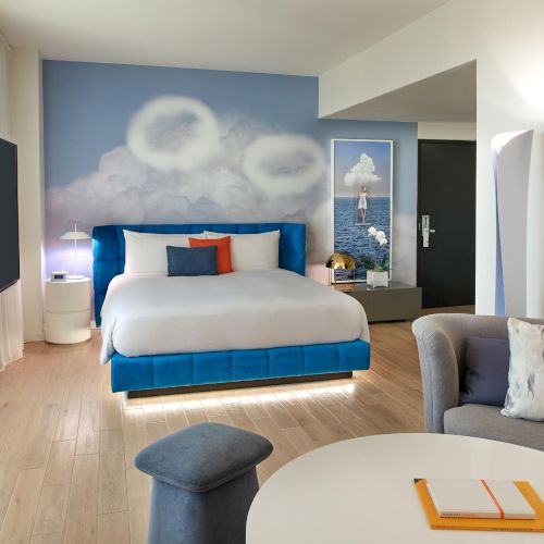 A bright hotel room with a blue-and-white color scheme: a large bed, wall mural of clouds, blue headboard, seating area, round table, and wall TV, ending with a period.