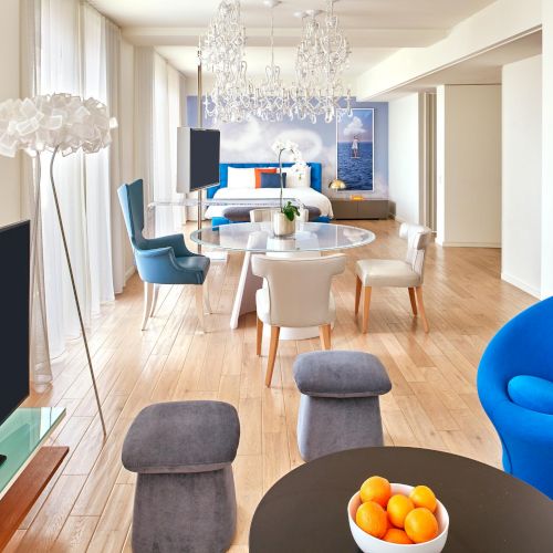 A bright, modern living room with a round black coffee table, orange fruit bowl, blue chairs, gray ottomans, a blue egg chair, and a dining area in the background.