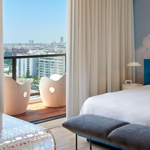 A modern hotel room with a balcony view of the city, white bed, blue accents, and a small seating area by sheer curtains, bright and airy.
