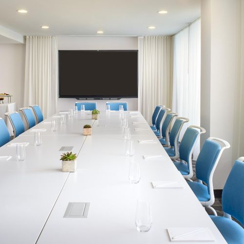 A bright, modern meeting room with a long white table, blue chairs, a wall-mounted screen, and a coffee station at the back, perfect for conferences.