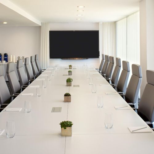 A modern conference room with a long white table, many gray chairs, plants, water glasses, and a large screen at the far end.