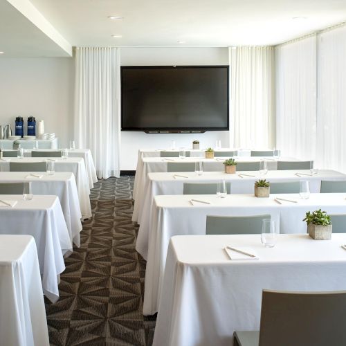 A bright conference room set with white tablecloths, rows of desks, a large screen at the front, and small plants on each table.