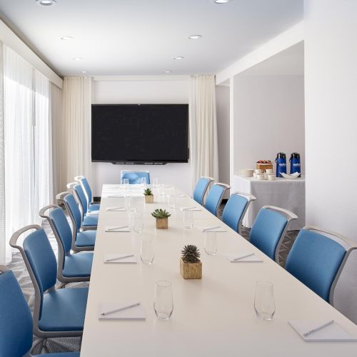 A sleek conference room with a long white table, blue chairs, a wall-mounted TV, and a refreshment station, set for a formal meeting.