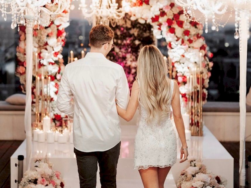 A couple walks hand in hand down a flower-adorned aisle under chandeliers at a wedding venue.