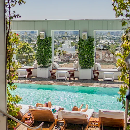 A poolside terrace with lounge chairs, a wooden deck, ivy-covered columns, and a city view in the background, bathed in sunny light.