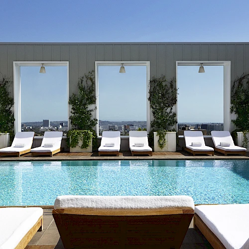 A rooftop pool scene with lounge chairs and tall potted plants along a row of windows, on a sunny day. 140 characters, ending with a period.