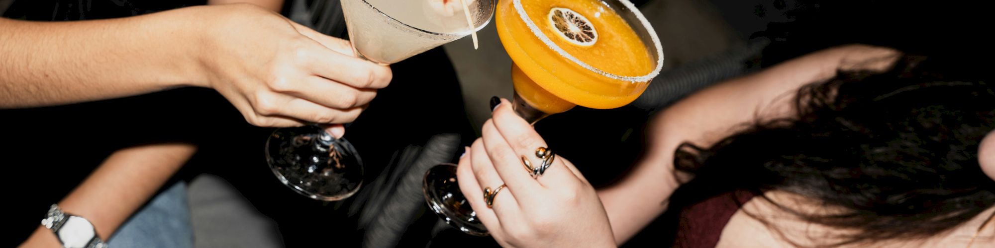 Two friends enjoy cocktails, one with a pale drink and the other with a bright orange beverage, while chatting and laughing.