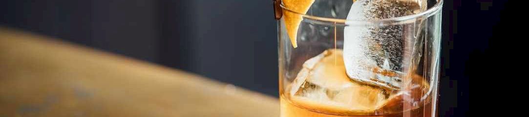 A glass of whiskey with ice, garnished with an orange twist and a small lemon rind, being poured over a wooden bar surface.