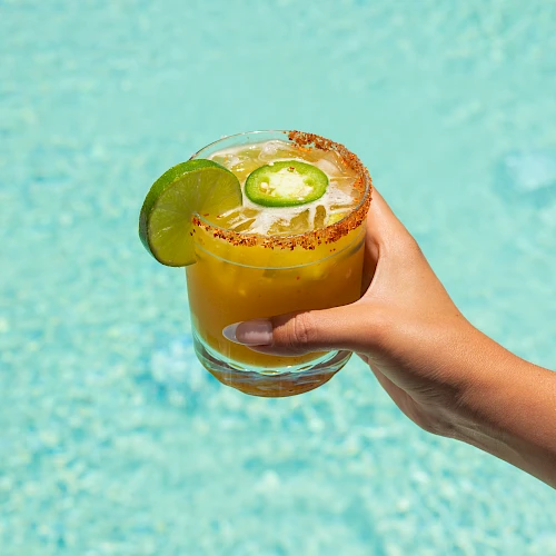 A hand holds a frosty cocktail with a lime wedge and chili salt rim, over a bright blue pool backdrop.