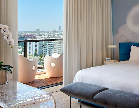 A chic hotel room with a balcony view: white bed, blue headboard, gray ottomans, sheer curtains, a small desk, and a bright cityscape beyond the balcony.
