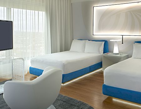 A bright hotel room with two single beds, white bedding and blue bases, a wall-lit headboard, modern chair, and a balcony view.