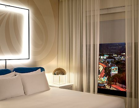 A modern hotel room with a lit geometric headboard, white bedding, a small bedside lamp, and a city view through a large window at night.