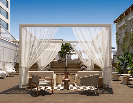 A modern rooftop lounge with white curtains, wicker sofas, and potted plants on a wooden deck under a clear blue sky.