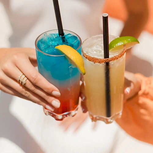 Two people clink colorful tropical cocktails with umbrellas, lime slices, and salted rims, in a sunny beachy vibe.