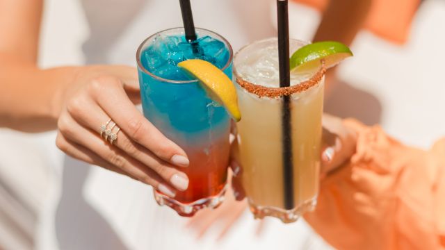 Two people clink colorful tropical cocktails with umbrellas, lime slices, and salted rims, in a sunny beachy vibe.