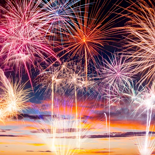 Colorful fireworks exploding above a sunset sky, bright trails and bursts lighting up the night.