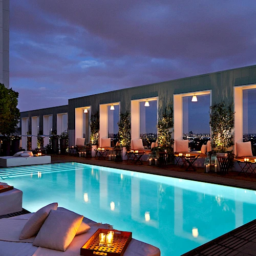 A luxurious rooftop pool at dusk with glowing water, stylish seating, warm lighting, and a modern building backdrop with large windows.