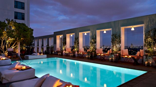 A luxurious rooftop pool at dusk with glowing water, stylish seating, warm lighting, and a modern building backdrop with large windows.