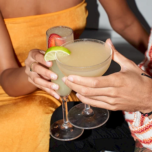 Two people clink margaritas, garnished with lime, in a sunny club vibe.