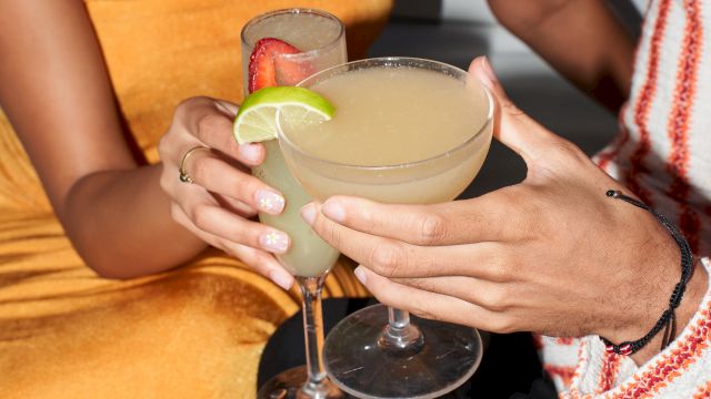 Two people clink margaritas, garnished with lime, in a sunny club vibe.