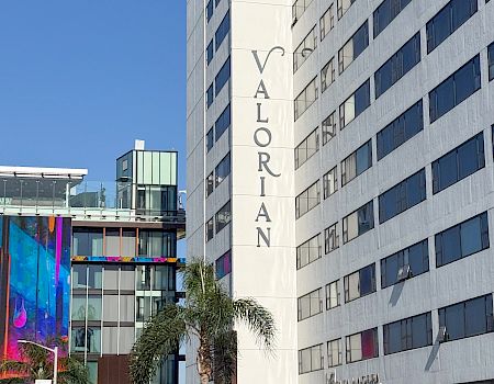 A tall white building with the word “VALORIAN” vertically on its side, a palm tree, and colorful glass structures nearby in an urban scene.