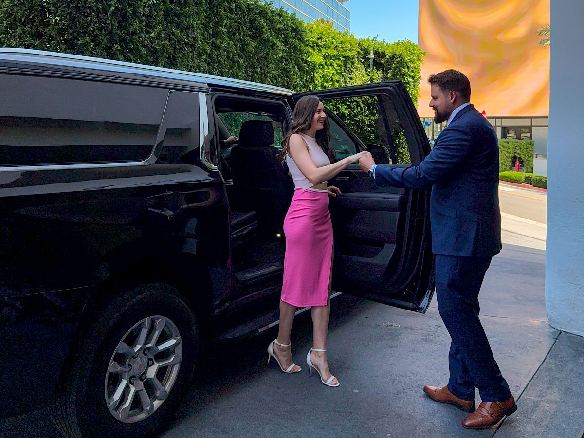 A stylish lady by a black SUV: woman in a pink dress exits while a man in a blue suit helps her, sunny outdoor setting.