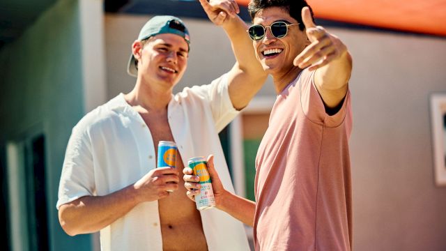 Two friends in sunny weather smile and toast with colorful canned drinks while one waves and wears sunglasses and the other a cap.