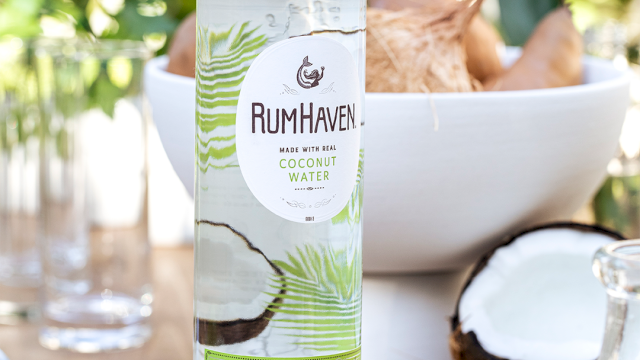 A RumHaven bottle labeled &ldquo;Made with real coconut water,&rdquo; with tropical drink ingredients on a sunny outdoor table.