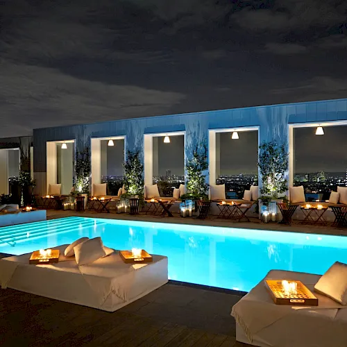 A nighttime rooftop resort pool with glowing lounge chairs and candles, flanked by doors and a city skyline beyond.