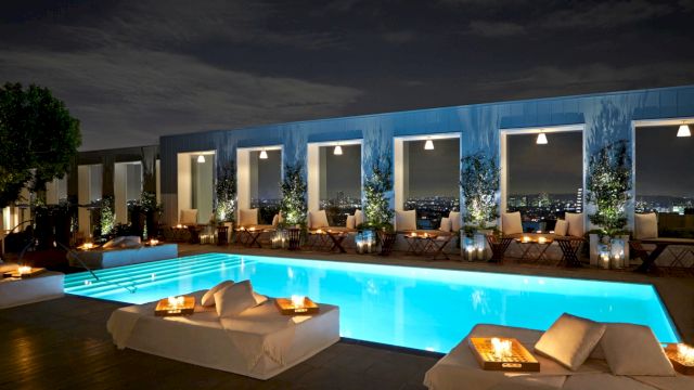A nighttime rooftop resort pool with glowing lounge chairs and candles, flanked by doors and a city skyline beyond.