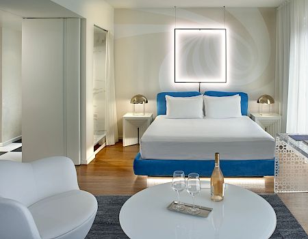 Modern bedroom: blue bed and white pillows, ceiling light frame, curtains, side tables, and a tray with glasses and a bottle.