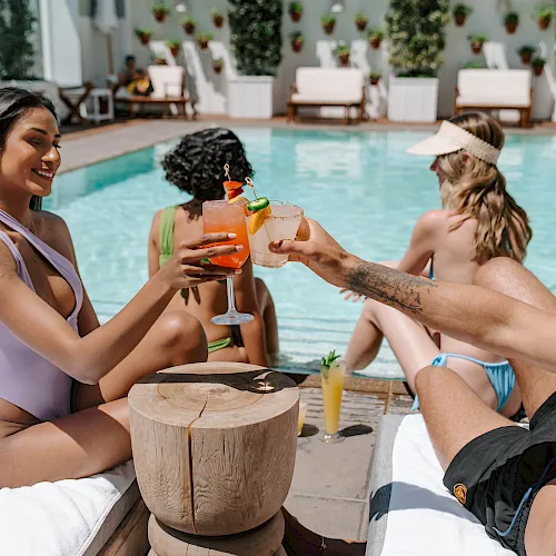 Friends clink tropical cocktails poolside on a sunny day, smiling and relaxing together.