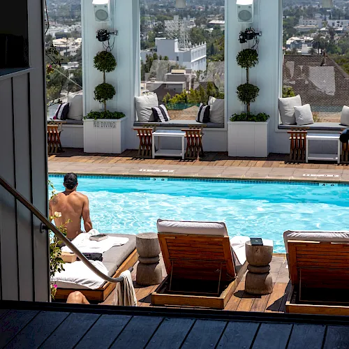 A sunny poolside patio with loungers, potted plants, and speakers, overlooking a cityscape from an upper terrace.