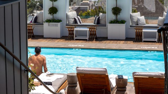 A sunny poolside patio with loungers, potted plants, and speakers, overlooking a cityscape from an upper terrace.