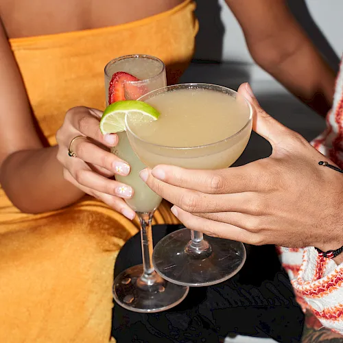 Two hands clink glasses of lime-garnished margarita-style cocktails at a sunny table.