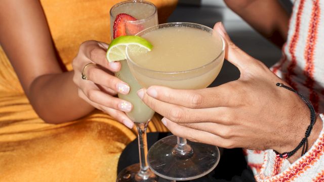 Two hands clink glasses of lime-garnished margarita-style cocktails at a sunny table.