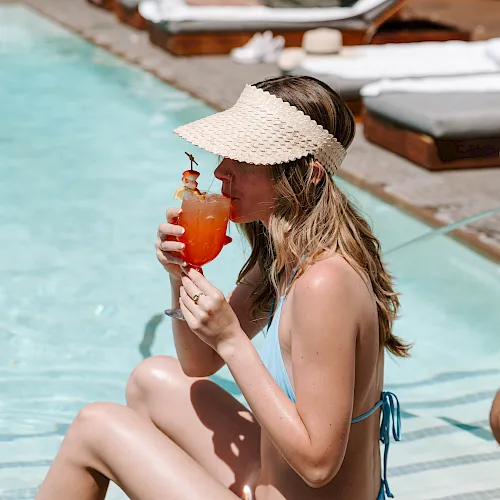 Woman lounges by a pool, sipping an orange cocktail with shrimp garnish under a straw hat.