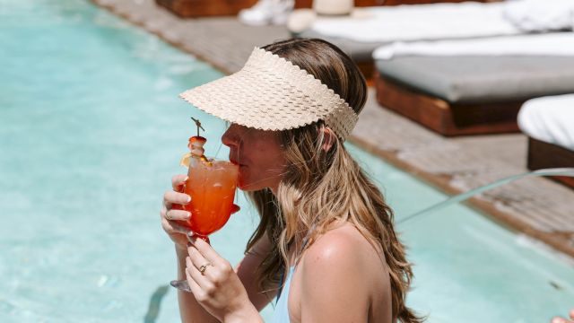 Woman lounges by a pool, sipping an orange cocktail with shrimp garnish under a straw hat.