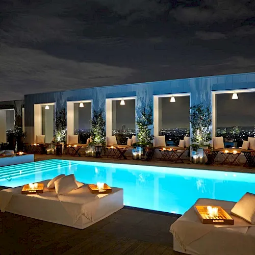 A rooftop infinity-style pool at dusk with sun loungers and city lights beyond large glass doors.
