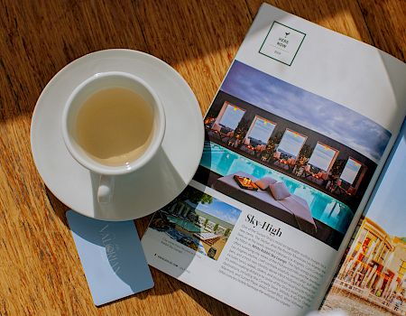A cup of tea sits beside an open travel magazine featuring a hotel/pool photo and “Sky-High” branding on the page.