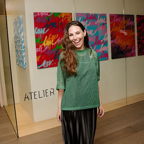 A woman in a green top smiles in an art gallery with colorful abstract paintings behind her.