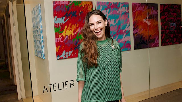 A woman in a green top smiles in an art gallery with colorful abstract paintings behind her.