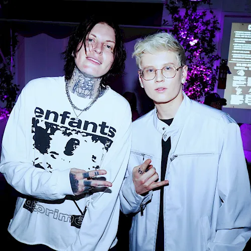 Two people in white lab coats pose at a neon-lit event, with a &ldquo;CAUTION&rdquo; display glowing in the background.