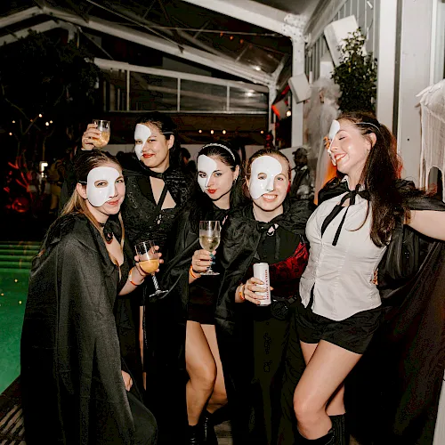 A group of friends in Halloween-style masks and capes cheers with drinks at a festive outdoor night party.