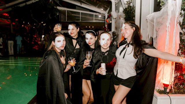 A group of friends in Halloween-style masks and capes cheers with drinks at a festive outdoor night party.