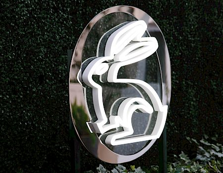 A shiny metal emblem with a bold white “L” logo, set against a wall of green leaves.