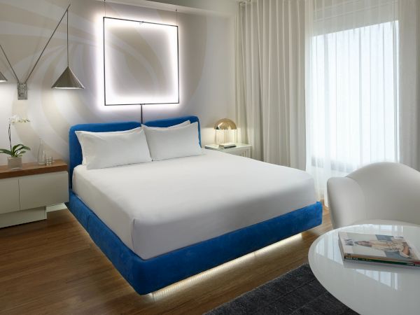 A bright modern bedroom with a blue upholstered bed, white pillows, hanging lamps, and a large window with sheer curtains.