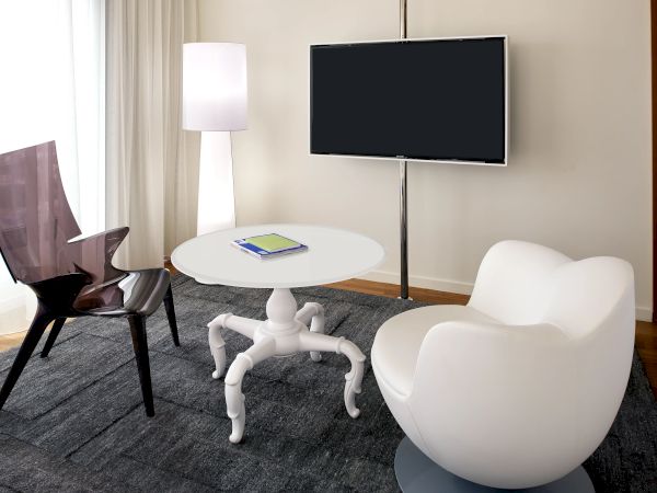 A bright meeting room with a round table, white chairs, a floor lamp, and a blank wall-mounted TV screen.