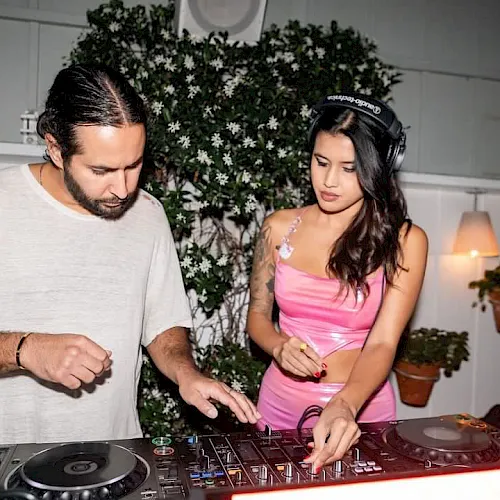 A man and woman in headphones mix music at a DJ console during a party with plants and lights behind them.